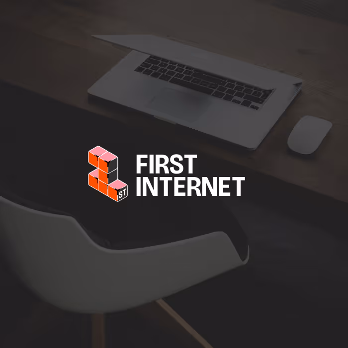 First Internet logo with stylized block letters next to a partially open laptop, wireless mouse, and modern chair on a wooden desk.